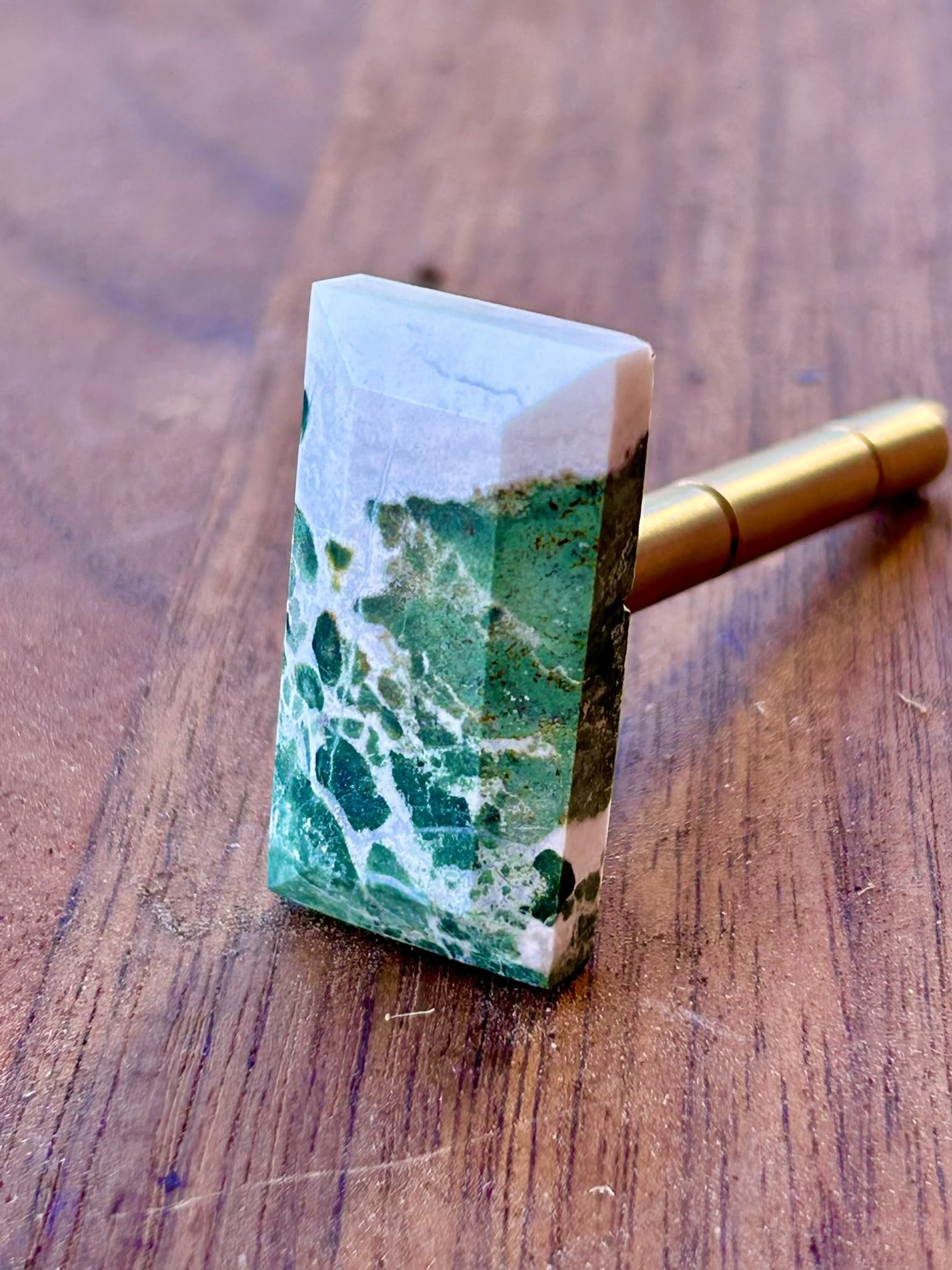 A moss agate, cut and polished, still on its brass dop stick on the bench: green dendrites in white chalcedony with a rind of matrix along one edge.