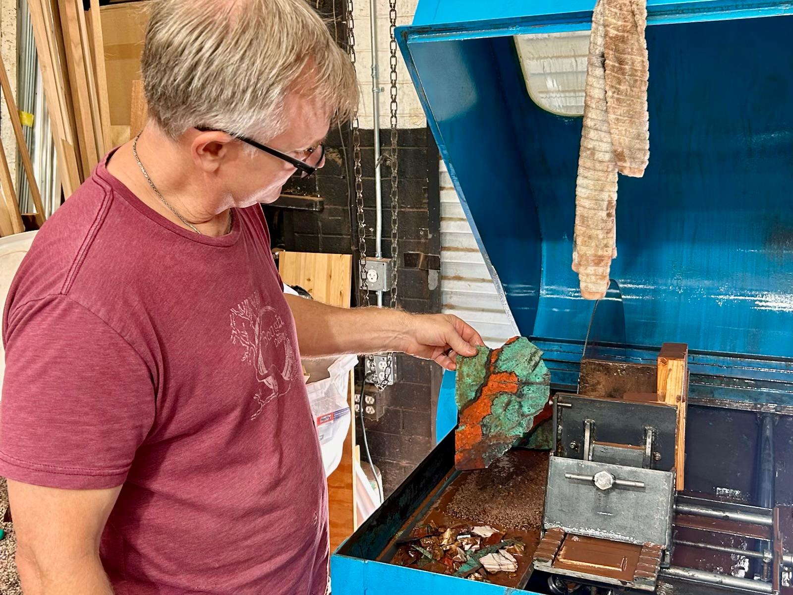 Jeremy at the slab saw holding a freshly cut slab, still wet, its face banded in blue green and rust orange.